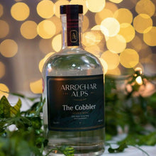 Load image into Gallery viewer, Clear glass bottle of Arrochar Alps The Cobbler gin with a dark green label and white writing. The bottle is sitting on a light coloured table with green foliage around it and circular, blurred fairy lights behind.
